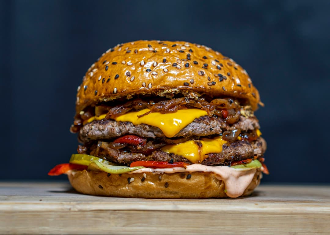 Hambúrguer de dois discos de carne com queijo derretido, cebola caramelizada e pão de brioche com gergelim, sobre uma tábua de madeira